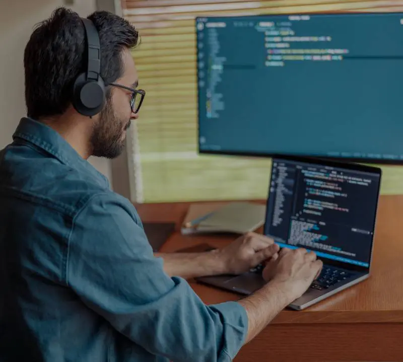A man coding on a laptop with a monitor displaying code in the background.